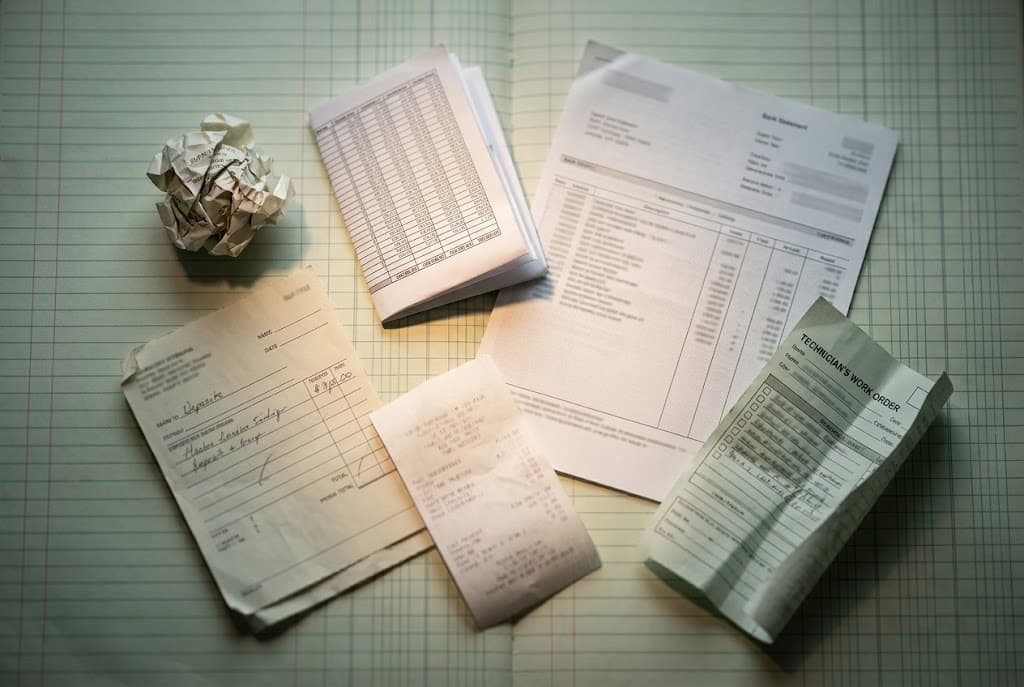 Six pieces of paper on a ledger pad: a crumpled supply-house receipt, a spreadsheet export, a bank statement, a deposit slip, a fuel receipt, and a technician's work order
